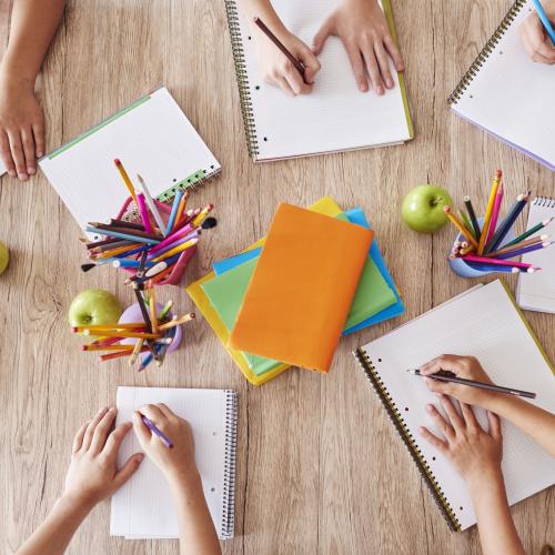 Prise de vue en plongée sur une table où sont installé des cahiers, des crayons et où l'on voit les mains en train d'écrire