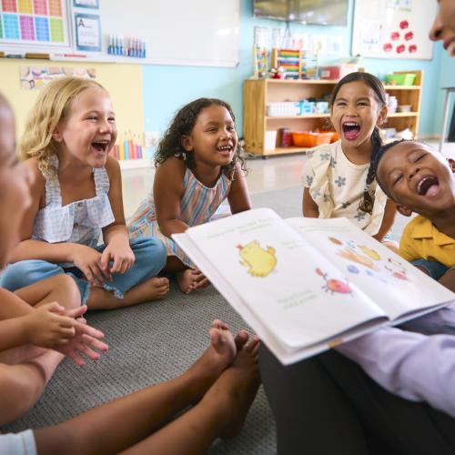 Des enfants souriants assis en cercle devant une femme qui tient un livre ouvert. 