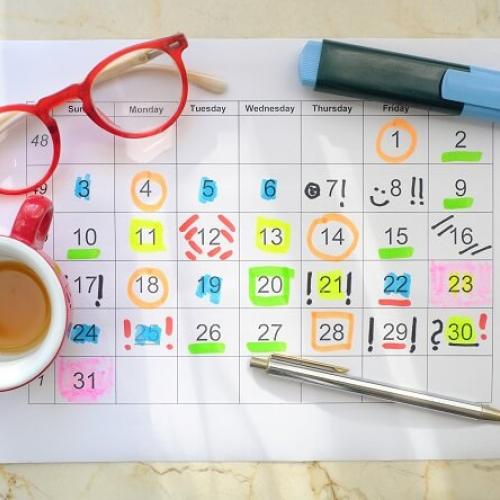 Sur une table, un planning dessiné sur une feuille, avec un tableau annoté et coloré au fluo. À côté, des lunettes, un tasse de thé ou de café, un bic et un stylo.