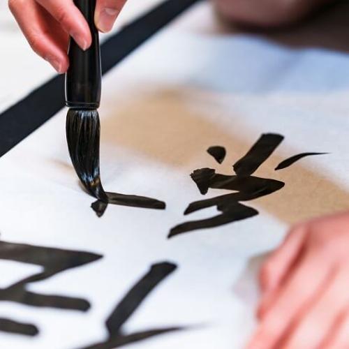 Gros plan sur des mains, dont une équipée d'un instrument pour la calligraphie, ainsi que des lettres en japonais en noir sur une feuille blanche