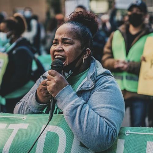 Une femme dans une manifestation, elle tient un micro dans lequel elle semble parler fort, on voit les pancartes devant et derrière elle.