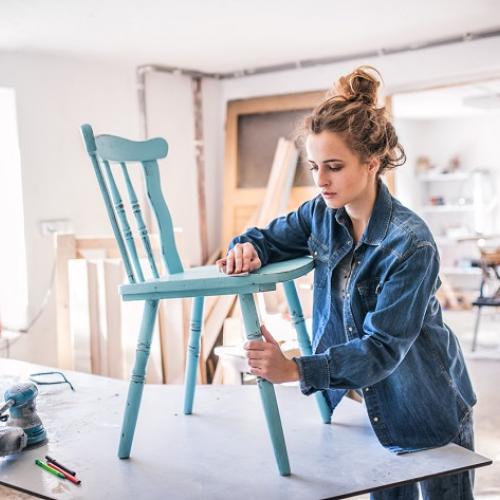 Une femme en bleu de travail ponce une chaise posée sur une table