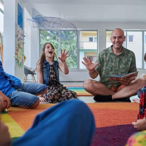 Des enfants assis par terre en cercle au tour d'un homme qui leur lit une histoire avec expression